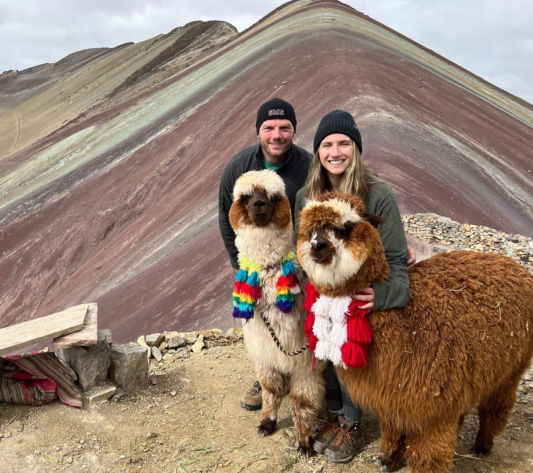 Male and female posed with two alpacas in front of rainbow mountain